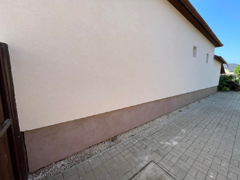 A ház oldala a kész, világos homlokzattal és barnás lábazattal, mellette térkő burkolatú udvar
