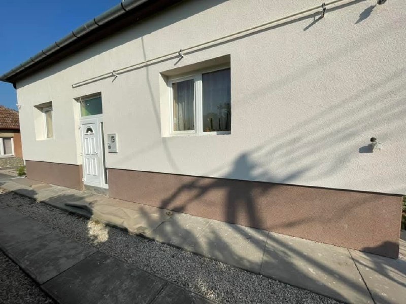 A ház homlokzata a bejárati ajtóval és ablakokkal, világos felülettel és barnás lábazattal